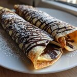 Two rolled Spiderweb Crepes drizzled with dark chocolate and dusted with powdered sugar on a white plate.