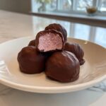 A stack of rich chocolate-covered Strawberry Truffles on a white plate, one cut open revealing the pink filling.