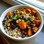 A close-up of a white bowl filled with Sweet Potato Black Bean Bowls, featuring brown rice, roasted sweet potatoes, black beans, corn, and cilantro.