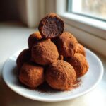 A stack of rich, cocoa-dusted Tiramisu Truffles piled on a white plate, one cut open showing the dark center.