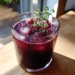 A close-up of a deep red Vampire Fig Thyme Punch served over ice in a sweating glass, garnished with fresh thyme sprigs.