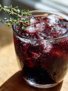 Close-up of a glass filled with dark red Vampire Fig Thyme Punch, ice, and garnished with fresh thyme.