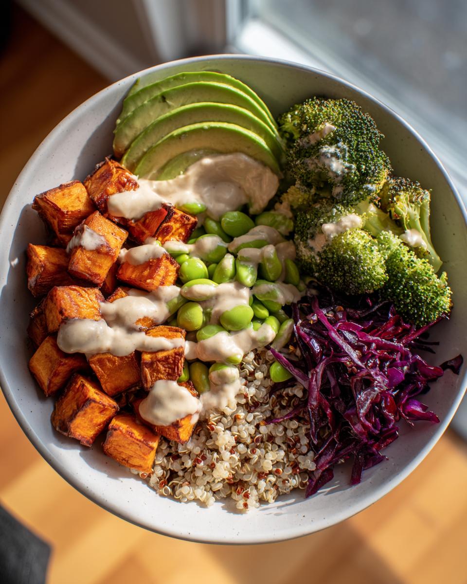 A vibrant Vegan Buddha Bowl featuring roasted sweet potatoes, avocado, edamame, broccoli, quinoa, and red cabbage, drizzled with tahini dressing.