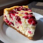 A close-up slice of White Chocolate Cranberry Cheesecake showing the creamy filling and bright red cranberries on a graham cracker crust.
