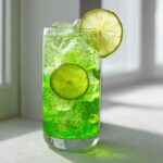 A tall glass filled with a bright green, fizzy Witch Brew Mocktail, ice, and lime slices.