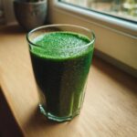 A tall glass filled with a thick, vibrant green Witchy Green Smoothie sitting on a wooden windowsill.