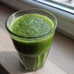 A glass filled with a vibrant Witchy Green Smoothie, topped with small bubbles and seeds, sitting on a wooden windowsill.