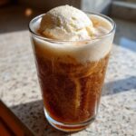 A tall glass containing a fizzy, dark brown Apple Cider Float topped with a scoop of melting vanilla ice cream.