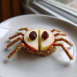 A fun Apple Spider Snack made from apple slices, pretzel legs, and raisin eyes, sitting on a white plate.
