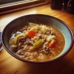 A steaming bowl of hearty Beef Cabbage Barley Soup filled with ground beef, barley, carrots, and cabbage on a wooden table.