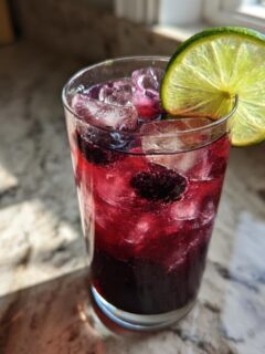 A tall glass filled with a vibrant, dark red Black Magic Mocktail, ice, and berries, garnished with a bright lime wheel.