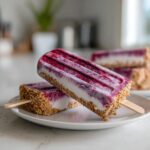 Two frozen Blackberry Cheesecake Pops resting on a white plate, showing the graham cracker crust and berry swirl.