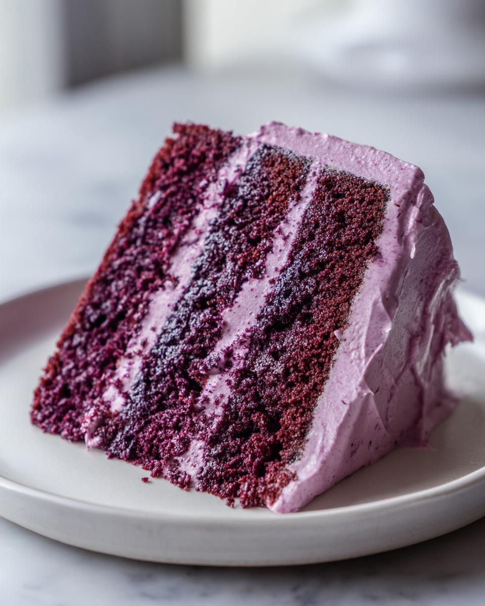 A close-up of a moist slice of Blackberry Velvet Cake showing deep purple layers and light purple blackberry frosting.