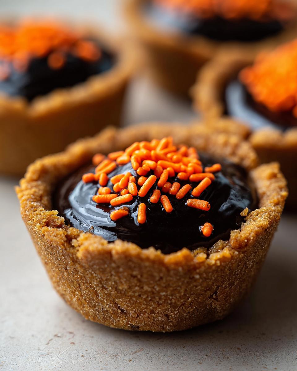 A close-up of a single Cauldron Cookie Cup filled with dark chocolate ganache and topped with bright orange sprinkles.
