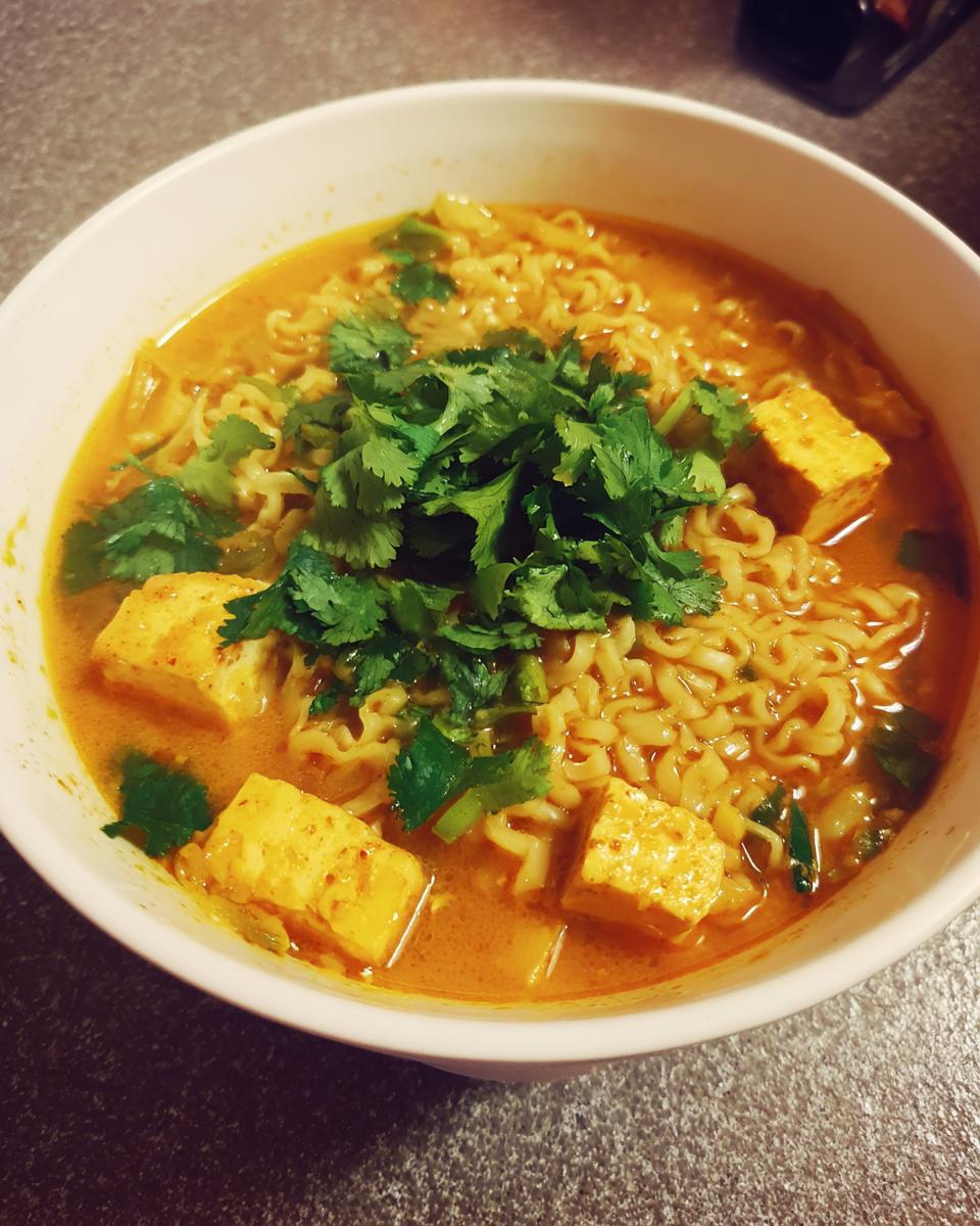 A close-up view of a white bowl filled with rich, orange Coconut Curry Ramen, topped with cubes of tofu and fresh cilantro.