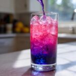 A stream of purple liquid is poured into a glass filled with ice, creating a vibrant color transition from deep blue to bright pink in the Color Changing Lemonade.