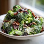 A close-up of a creamy Broccoli Bacon Salad featuring fresh broccoli florets coated in dressing, mixed with crispy bacon, raisins, and sunflower seeds.