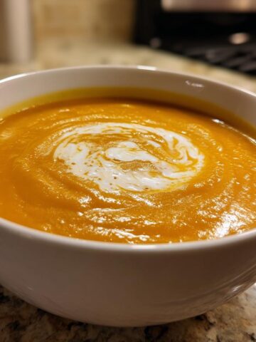 A close-up of a white bowl filled with vibrant orange Pumpkin Soup, topped with a swirl of cream.