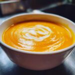 Close-up of a vibrant orange bowl of smooth Pumpkin Soup, topped with a swirl of white cream.