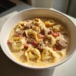 Close-up of a white bowl filled with Creamy Tortellini Soup With Sausage, topped with black pepper.