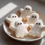 Five fluffy, white Ghost Cookies coated in sugar, arranged on a white plate with black candy eyes.