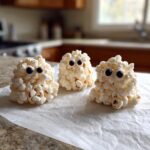 Three adorable Ghost Popcorn Balls made from popcorn and decorated with black candy eyes, sitting on parchment paper.