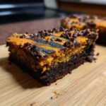 A close-up of a rich, fudgy Halloween Brookies slice showing a dark brownie base and a marbled orange and black topping sprinkled with mini chocolate chips.