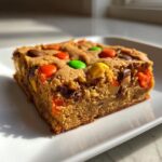 A thick, chewy square of Halloween Candy Cookie Bars studded with colorful M&Ms and peanut butter candies, sitting on a white plate.