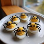 Six spooky Halloween Deviled Eggs decorated to look like spiders with black legs and eyes on a white plate.