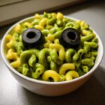 Bowl of Halloween Monster Mac And Cheese featuring green and yellow pasta with two black olive eyes.