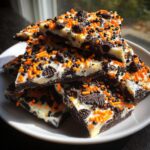 A stack of triangular pieces of Halloween Oreo Bark featuring a dark chocolate base, white chocolate topping, orange and black sprinkles, and crushed cookies.