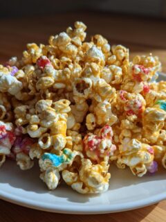 A mound of golden caramel Hocus Pocus Popcorn mixed with colorful candy pieces served on a white plate.