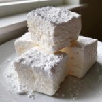 Close-up of three fluffy, homemade Stay Puft Marshmallows stacked on a white plate, heavily dusted with powdered sugar.
