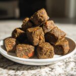 A close-up stack of dark brown, dense, cubed Werewolf Kibble treats with visible orange flecks, served on a white plate.