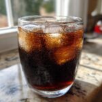 Close-up of a sweating glass filled with dark, bubbly Black Lemonade and ice cubes.