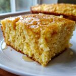Close-up of a square slice of moist Maple Cornbread drizzled generously with golden syrup on a white plate.