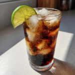 A tall glass filled with ice and dark soda, garnished with a bright lime wedge, representing a Midnight Malibu Dirty Soda.