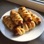 A plate of freshly baked Mummy Dogs wrapped in pastry strips with olive eyes, ready to eat.