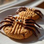 Two decorated Peanut Butter Spider Cookies on a white plate, featuring candy eyes and piped chocolate legs.