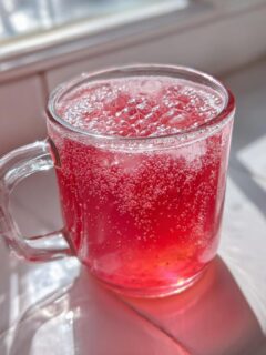 A clear glass mug filled with bright red, fizzy Pink Witch Punch sitting on a sunlit white windowsill.