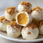 A stack of white chocolate-covered Pumpkin Cheesecake Truffles, one cut in half revealing the orange filling and topped with cookie crumbs.