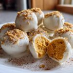 A pile of white chocolate covered Pumpkin Cheesecake Truffles, one cut open showing the orange filling and topped with graham cracker crumbs.