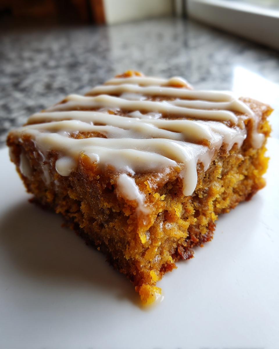 A close-up, slightly angled shot of a moist, orange-hued Pumpkin Spice Latte Bars square topped with a white vanilla glaze drizzle.
