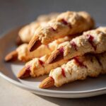 Close-up of several Witch Finger Cookies stacked on a plate, featuring almond slivers as nails and red jam for a bloody effect.