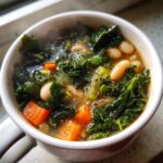 Close-up of a steaming white mug filled with vibrant White Bean Kale Soup, featuring dark green kale, white beans, and bright orange carrots.