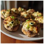 Several baked Stuffed Mushroom Eyeballs garnished with green olives and scallions on a white platter.