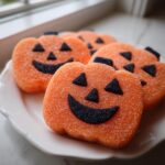 Several bright orange, sugar-coated Jack O Lantern Cookies with black sprinkle faces sitting on a white scalloped plate.