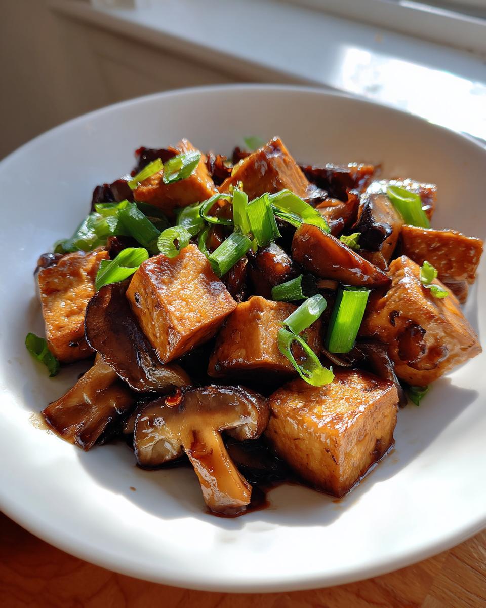Close-up of glossy, saucy cubes of tofu and sliced mushrooms in a Tofu Mushroom Stir Fry, garnished with green onions.