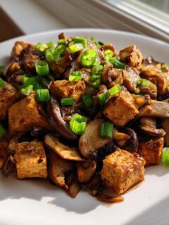 A close-up of a white plate holding a serving of Tofu Mushroom Stir Fry, topped generously with bright green chopped scallions.