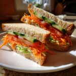 Two halves of the Ultimate Veggie Sandwich stacked with shredded carrots, lettuce, tomato, and cucumber on whole wheat bread.
